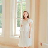 A girl wearing the KIKI and JOJO Girls Pearl-Embroidered Satin Dress in Pure White stands indoors by tall windows, bathed in natural light.