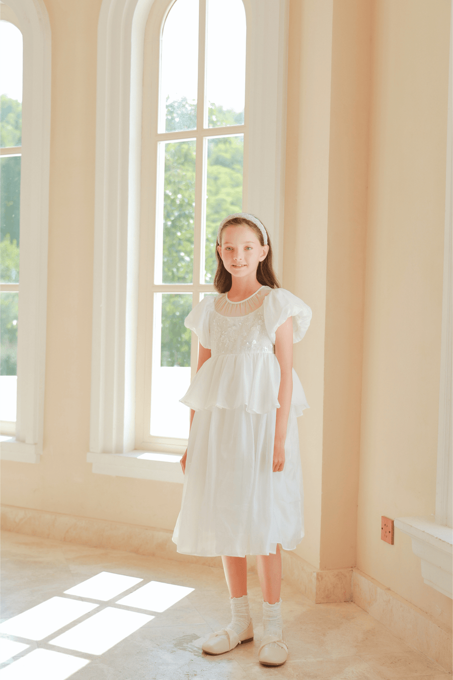 A girl wearing the KIKI and JOJO Girls Pearl-Embroidered Satin Dress in Pure White stands indoors by tall windows, bathed in natural light.