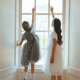 Two young girls stand by a large window; one wears the KIKI and JOJO Girls Pearl-Embroidered Satin Dress in Pure White and reaches up toward the top of the window while the other girl looks on.