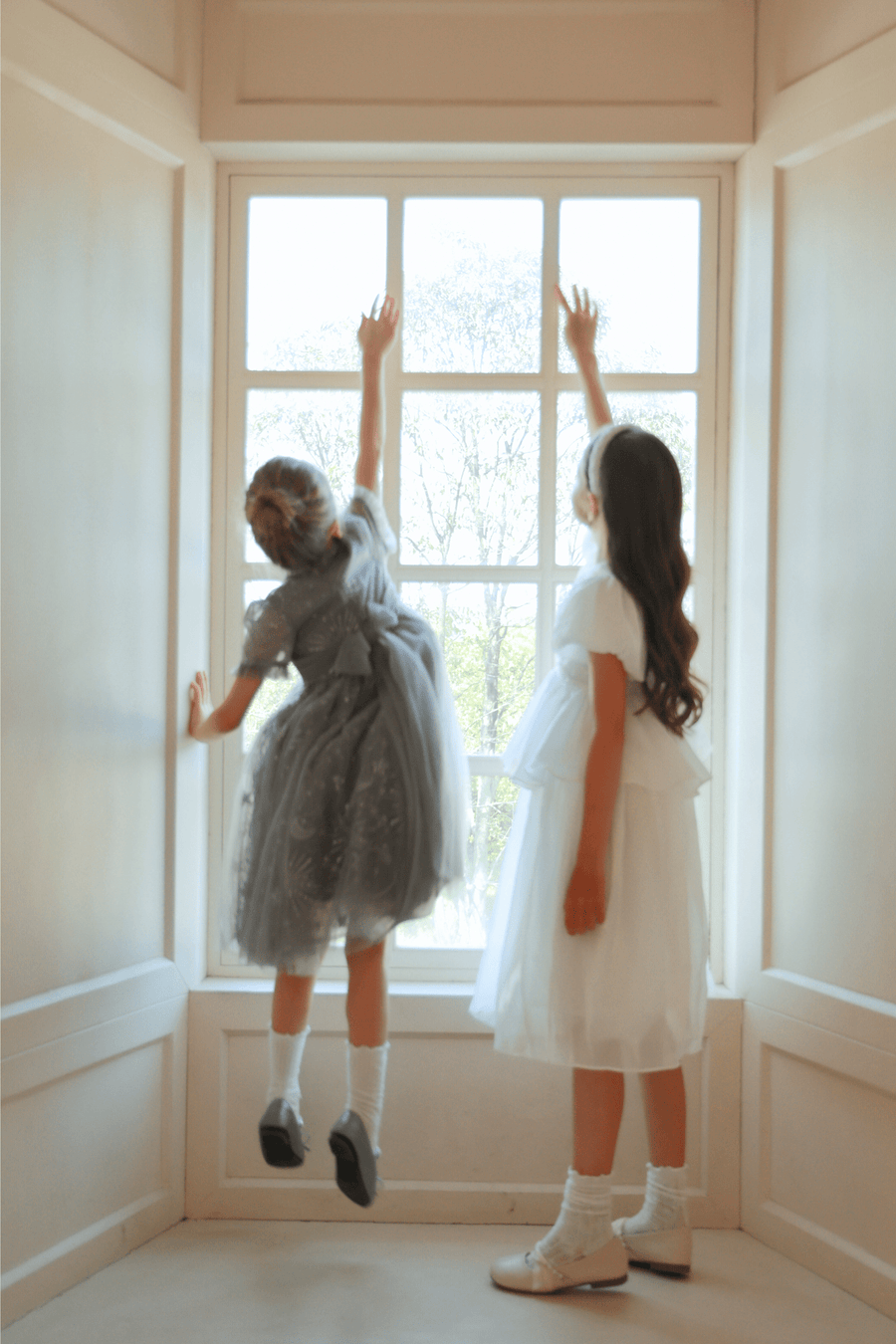 Two young girls stand by a large window; one wears the KIKI and JOJO Girls Pearl-Embroidered Satin Dress in Pure White and reaches up toward the top of the window while the other girl looks on.