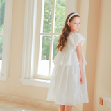 A girl wears the KIKI and JOJO Girls Pearl-Embroidered Satin Dress in Pure White, standing indoors near large windows and looking back over her shoulder as sunlight streams in.