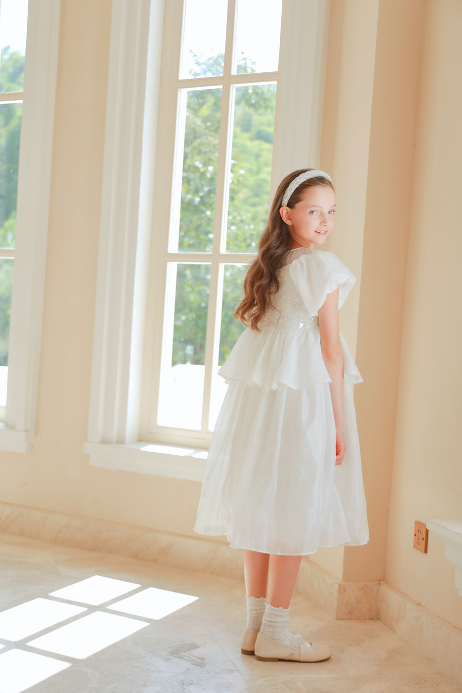 A girl wears the KIKI and JOJO Girls Pearl-Embroidered Satin Dress in Pure White, standing indoors near large windows and looking back over her shoulder as sunlight streams in.
