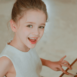 A young girl in the KIKI and JOJO Girls Pearlescent Elegance Tweed Dress in Soft Champagne smiles while playing piano, looking up at the camera.