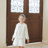 A young girl beams in front of a grand wooden door, elegantly dressed in the KIKI and JOJO Girls Pearlescent Elegance Tweed Dress in Soft Champagne, perfect for special occasions.