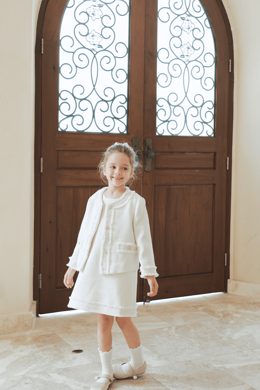 A young girl beams in front of a grand wooden door, elegantly dressed in the KIKI and JOJO Girls Pearlescent Elegance Tweed Dress in Soft Champagne, perfect for special occasions.