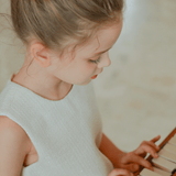 A young girl in the KIKI and JOJO Girls Pearlescent Elegance Tweed Dress in Soft Champagne plays the piano, gazing down at the keys with both hands on the keyboard.