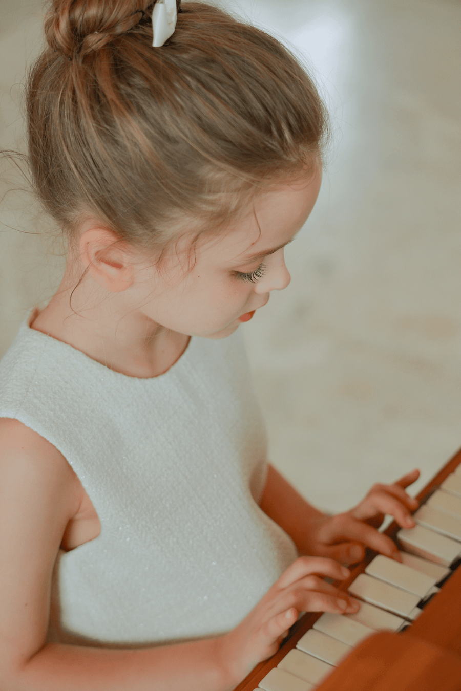 A young girl in the KIKI and JOJO Girls Pearlescent Elegance Tweed Dress in Soft Champagne plays the piano, gazing down at the keys with both hands on the keyboard.
