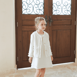 A young girl wears the KIKI and JOJO Girls Pearlescent Elegance Tweed Dress in Soft Champagne, standing indoors on a stone floor before grand wooden doors with ornate ironwork.