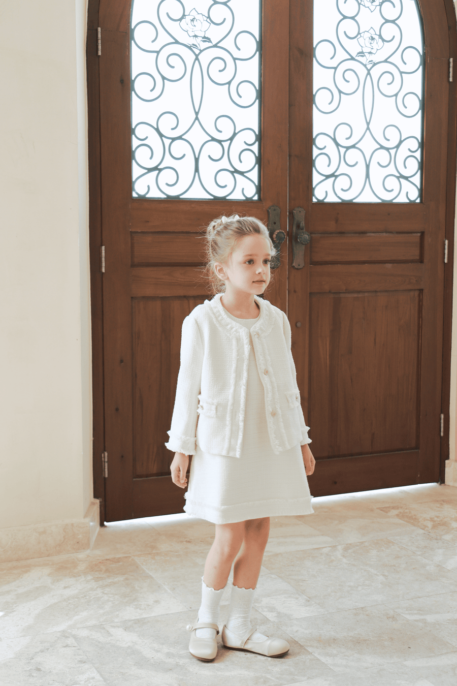 A young girl wears the KIKI and JOJO Girls Pearlescent Elegance Tweed Dress in Soft Champagne, standing indoors on a stone floor before grand wooden doors with ornate ironwork.