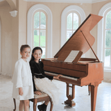 Two young girls in formal attire pose by a grand piano; one sits at the keys wearing the KIKI and JOJO Girls Pearlescent Elegance Tweed Dress in Soft Champagne, while the other stands beside her in a bright room with arched windows.