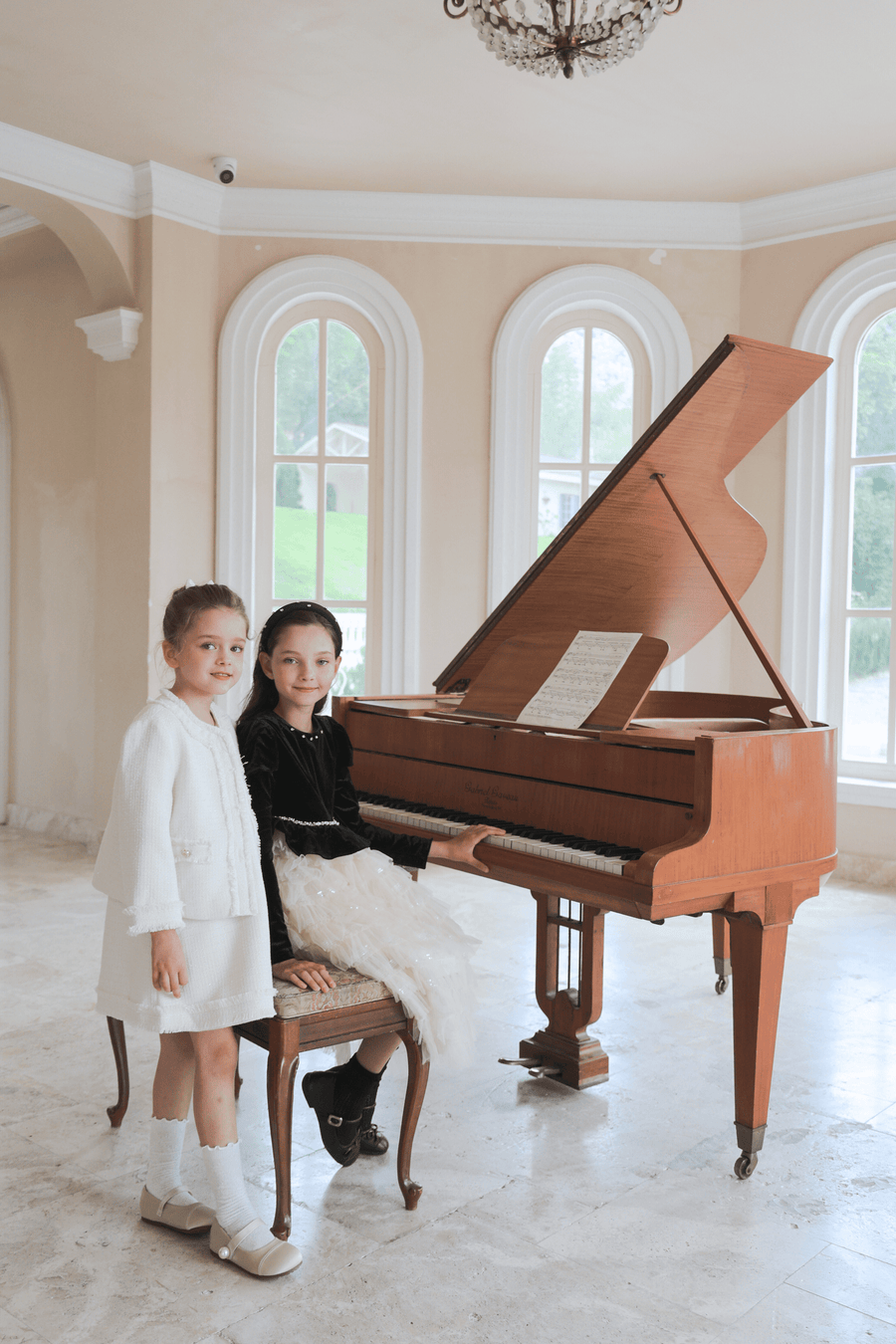 Two young girls in formal attire pose by a grand piano; one sits at the keys wearing the KIKI and JOJO Girls Pearlescent Elegance Tweed Dress in Soft Champagne, while the other stands beside her in a bright room with arched windows.