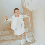 A young girl smiles on a curved staircase, wearing the KIKI and JOJO Girls Pearlescent Elegance Tweed Dress in Soft Champagne.