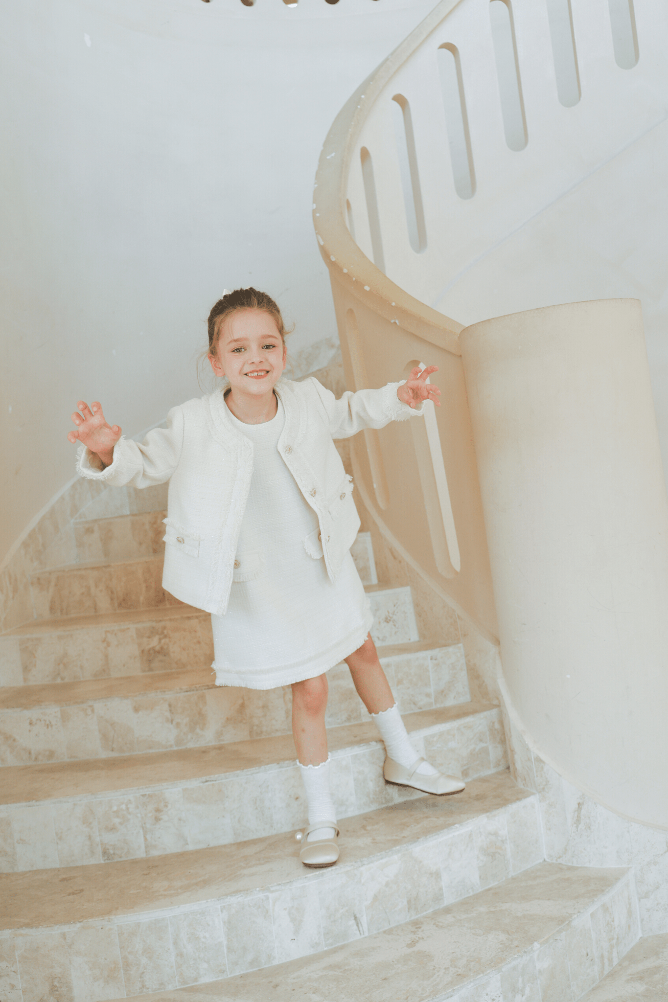 A young girl smiles on a curved staircase, wearing the KIKI and JOJO Girls Pearlescent Elegance Tweed Dress in Soft Champagne.