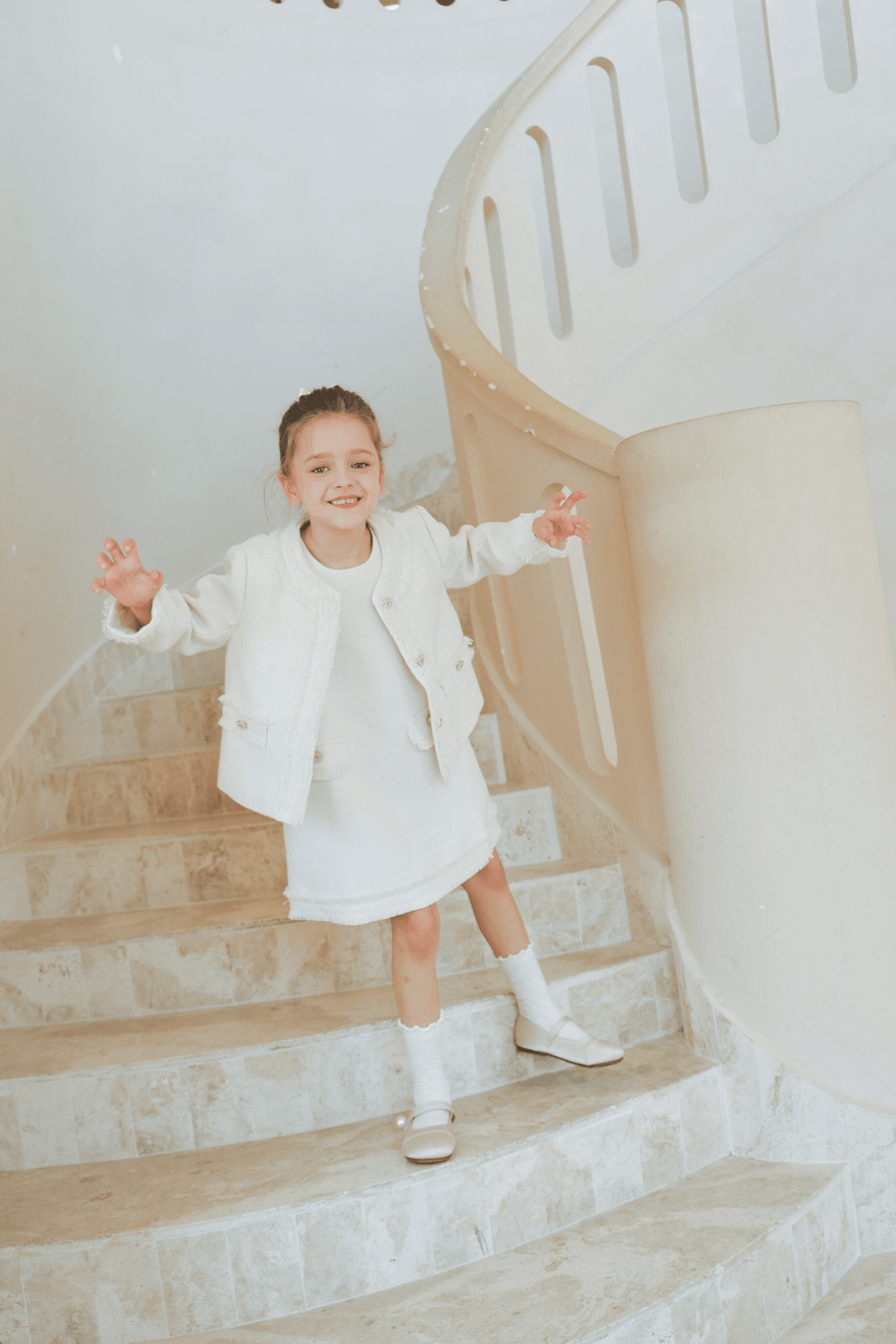 A young girl smiles on a curved staircase, wearing the KIKI and JOJO Girls Pearlescent Elegance Tweed Dress in Soft Champagne.