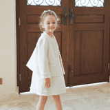 A young girl smiles indoors in front of ornate wooden doors, wearing the KIKI and JOJO Girls Pearlescent Elegance Tweed Dress in Soft Champagne—perfect for any elegant outing.
