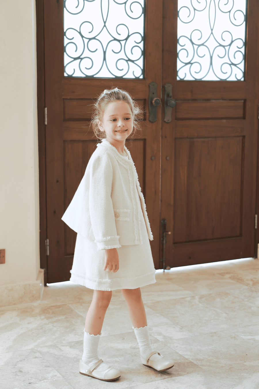 A young girl smiles indoors in front of ornate wooden doors, wearing the KIKI and JOJO Girls Pearlescent Elegance Tweed Dress in Soft Champagne—perfect for any elegant outing.