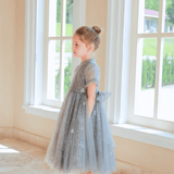 A young girl wearing KIKI and JOJO’s Girls Plated Metal Bow Hair Clips (2 Pack) in silver stands indoors by large windows, gazing outside with her hands in her pockets.