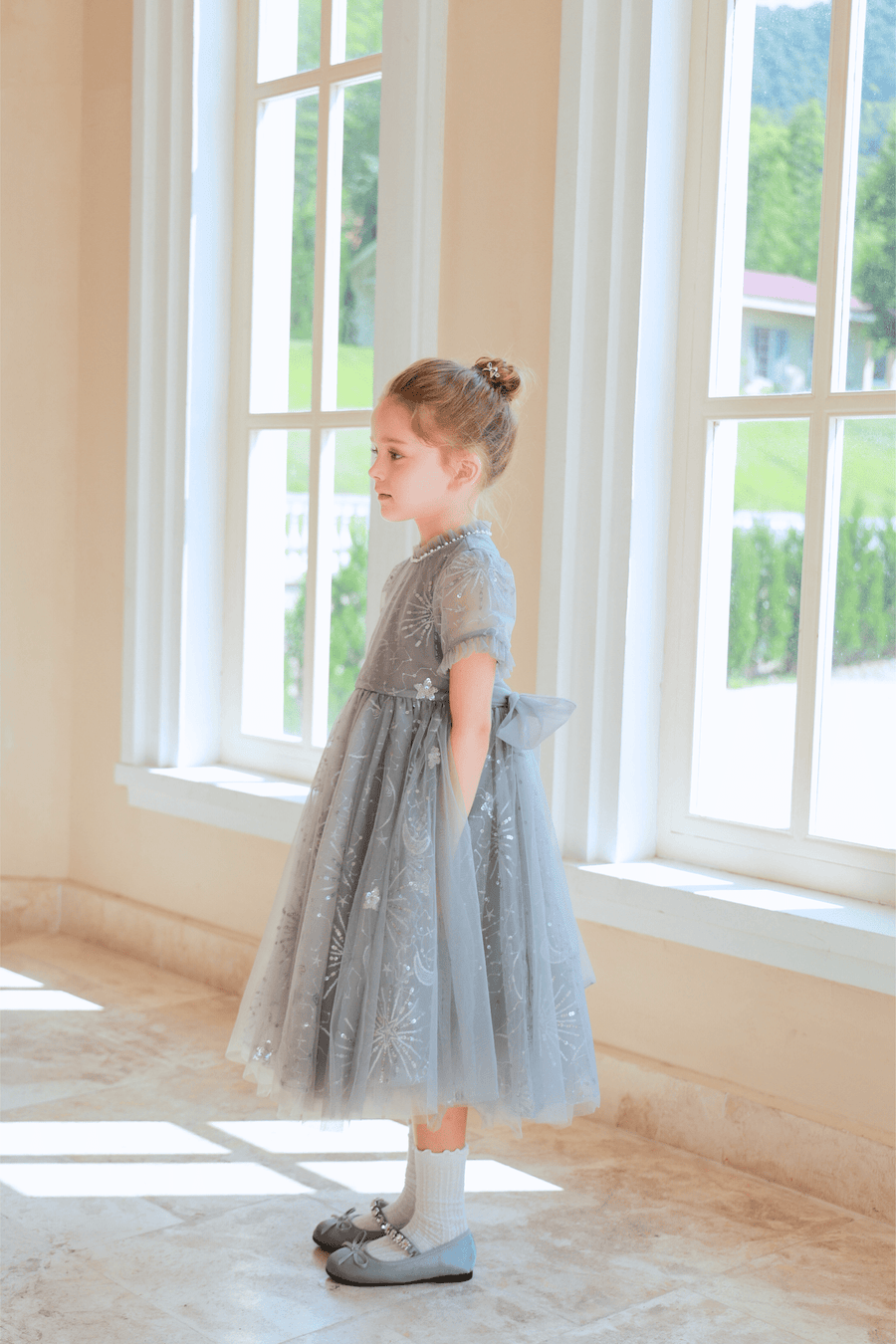 A young girl wearing KIKI and JOJO’s Girls Plated Metal Bow Hair Clips (2 Pack) in silver stands indoors by large windows, gazing outside with her hands in her pockets.
