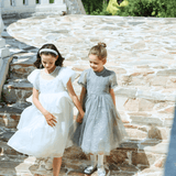Two young girls in formal dresses wear KIKI and JOJO Girls Plated Metal Bow Hair Clips (2 Pack) - Silver as they walk hand-in-hand down stone steps outside, with greenery and a white railing behind them.