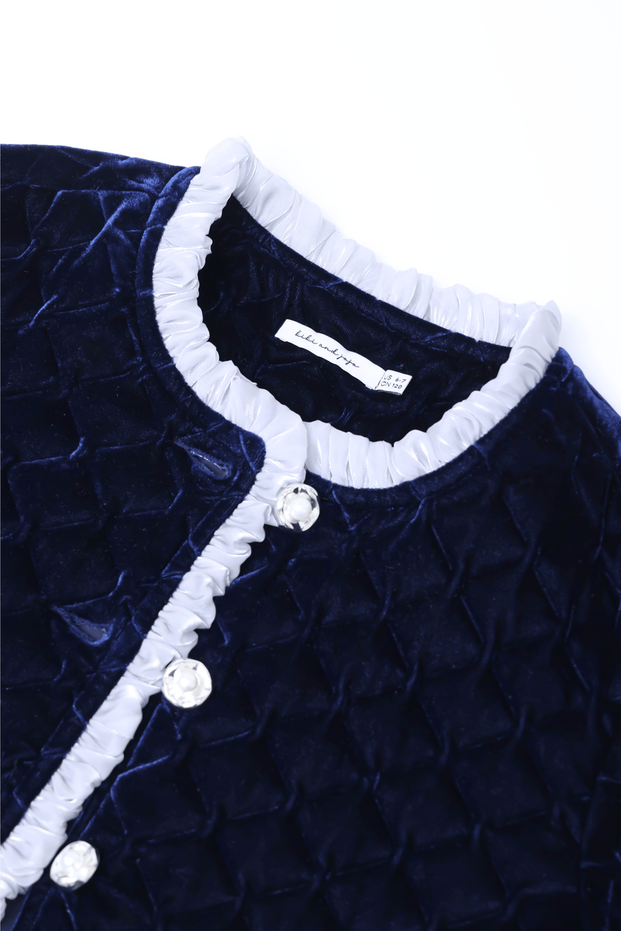 Close-up of the KIKI and JOJO Girls Quilted Ruffle Jacket in Sapphire Blue, featuring diamond-quilted velvet, clear petal-shaped buttons, and a light gray ruffled collar trim.