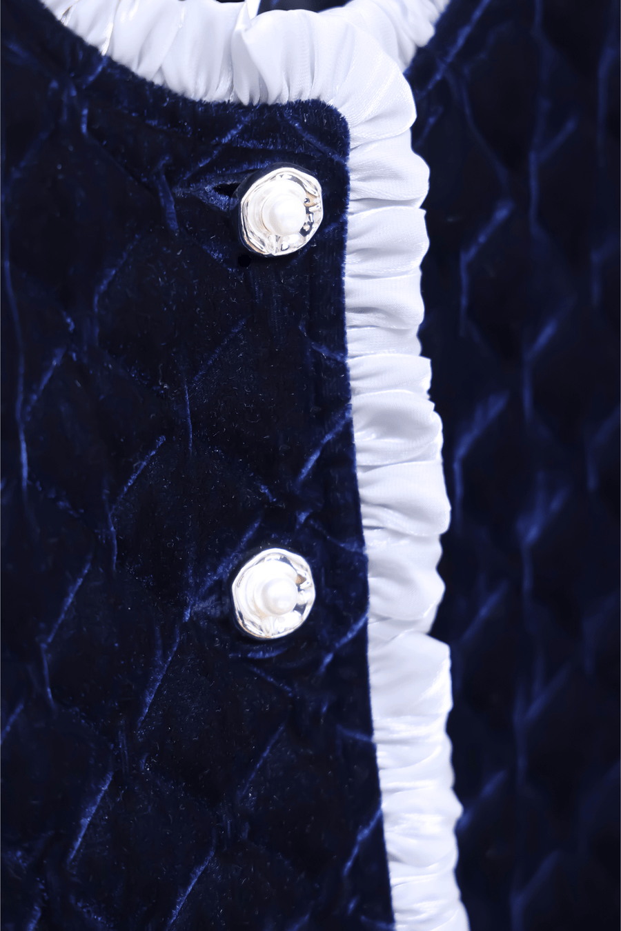 Close-up of the Girls Quilted Ruffle Jacket in Sapphire Blue by KIKI and JOJO, showcasing diamond-quilted velvet, two petal-shaped buttons, and a white ruffled edge trim.