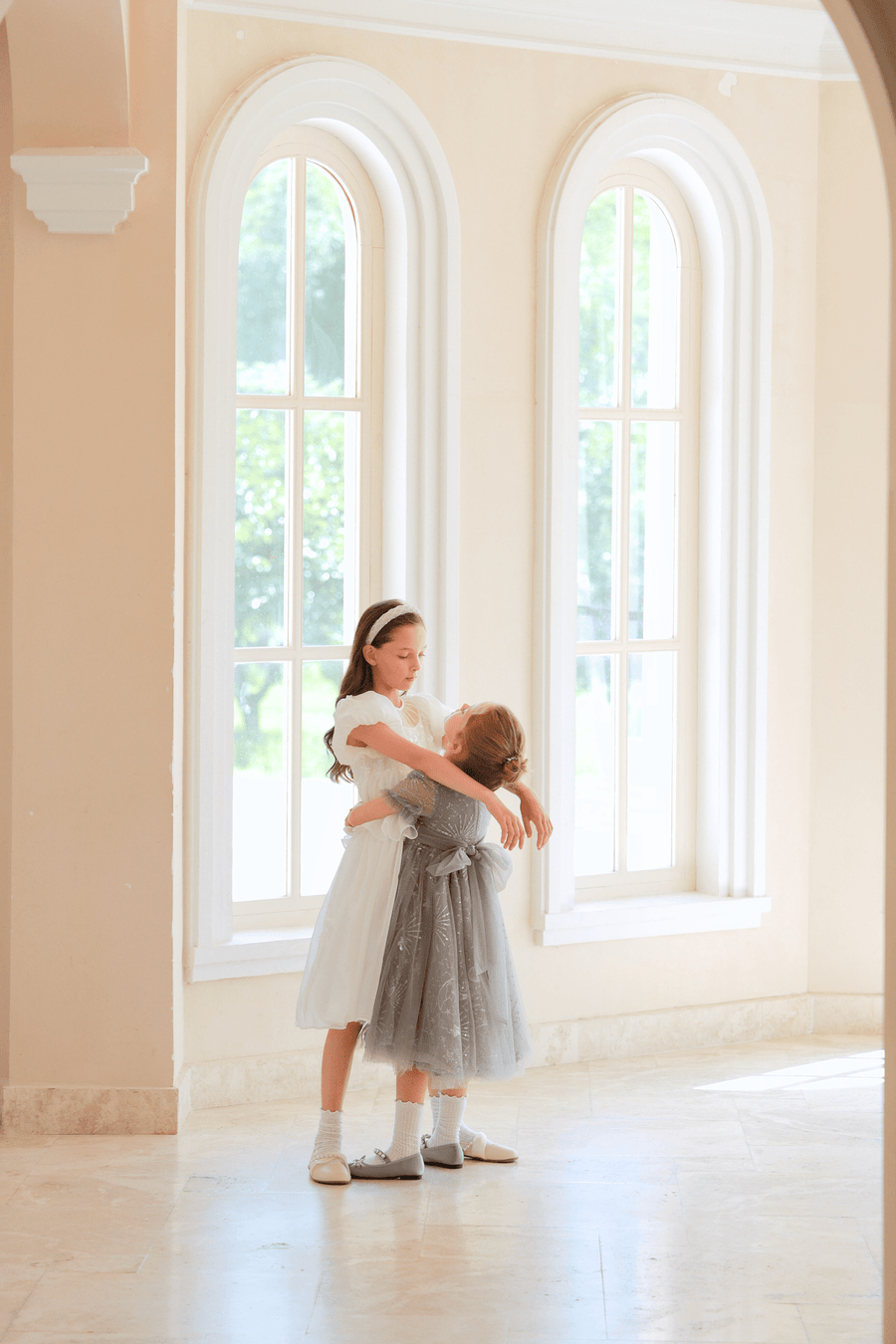 Two young girls, one wearing the KIKI and JOJO Girls Sequin Embroidered Tulle Dress in Starlight Silver, stand embracing before tall arched windows in a bright, sunlit room.