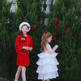 Two young girls stand before tall green bushes; one wears the KIKI and JOJO Girls Tweed Dress in cranberry red with an A-line silhouette and a white beret, while the other dons a white ruffled dress. Both have long hair and face different directions.