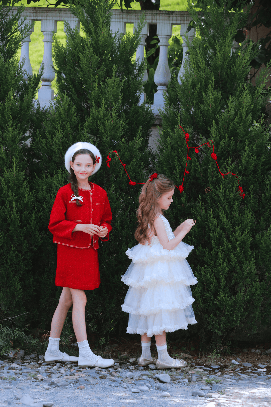 Two young girls stand before tall green bushes; one wears the KIKI and JOJO Girls Tweed Dress in cranberry red with an A-line silhouette and a white beret, while the other dons a white ruffled dress. Both have long hair and face different directions.