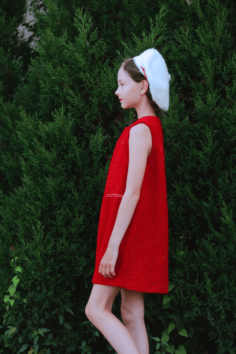 A young person in the KIKI and JOJO Girls Tweed Dress in cranberry red and a white beret stands in profile against green bushes.