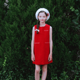 A young girl wears the KIKI and JOJO Girls Tweed Dress in cranberry red and a white beret, standing before dense green bushes and looking at the camera with a neutral expression.
