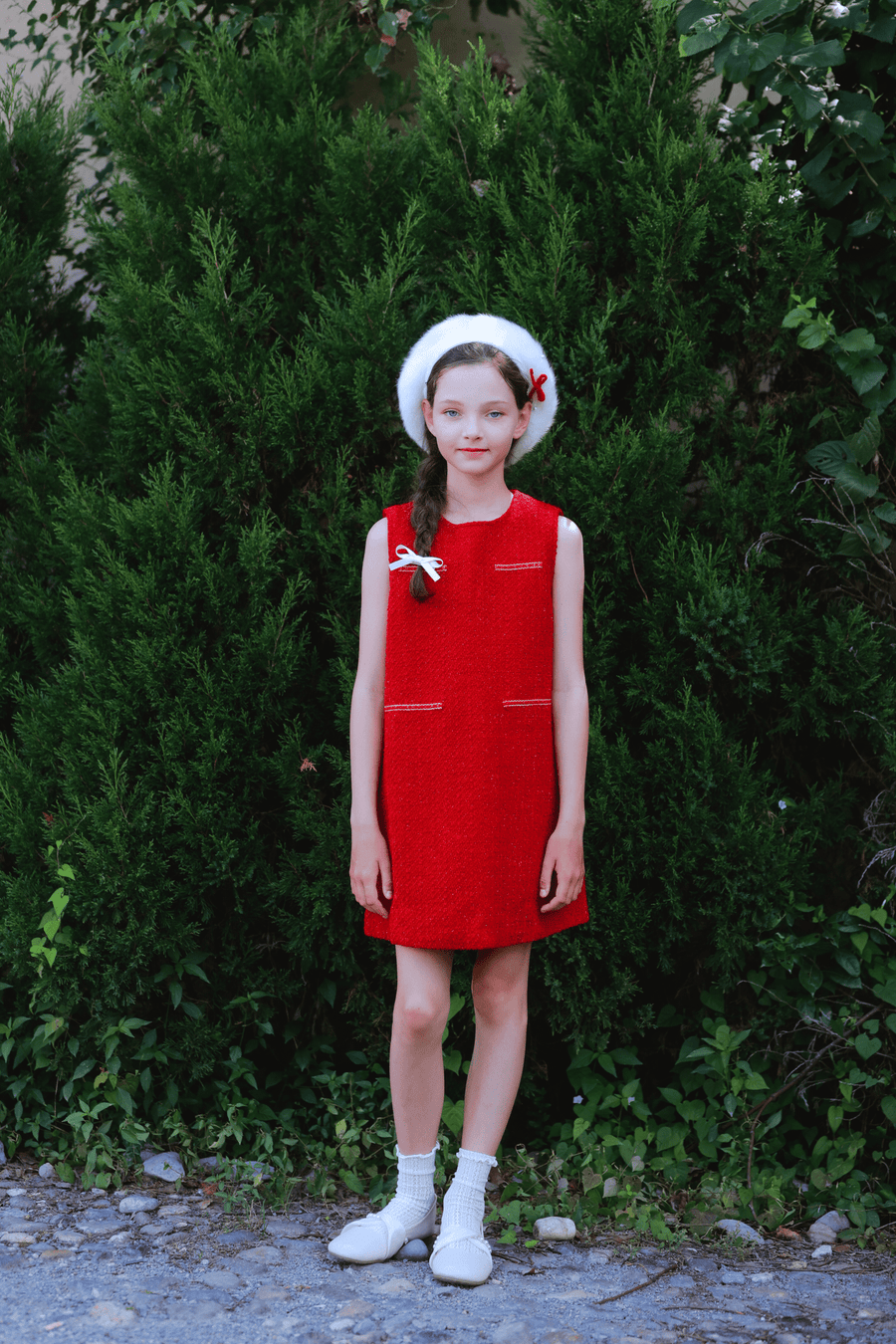 A young girl wears the KIKI and JOJO Girls Tweed Dress in cranberry red and a white beret, standing before dense green bushes and looking at the camera with a neutral expression.