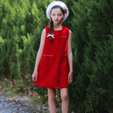 A girl in a KIKI and JOJO Girls Tweed Dress - Cranberry Red, paired with white socks and a white hat, stands on a cobblestone path by tall green bushes—ideal festive attire.