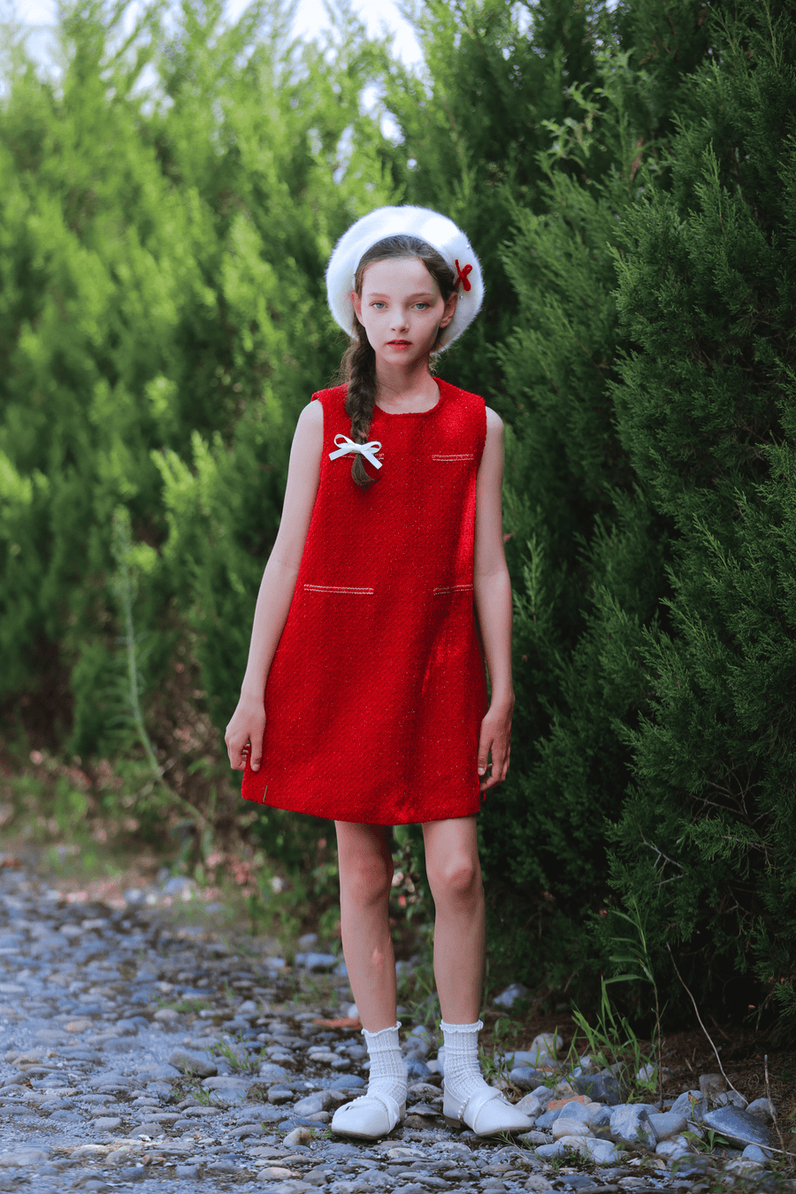A girl in a KIKI and JOJO Girls Tweed Dress - Cranberry Red, paired with white socks and a white hat, stands on a cobblestone path by tall green bushes—ideal festive attire.