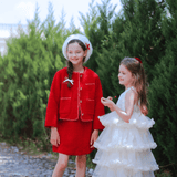 Two young girls stand outdoors; one wears the KIKI and JOJO Girls Tweed Dress in Cranberry Red with a beret, while the other is in a white ruffled dress. Lush green foliage provides a festive backdrop.
