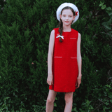 A young girl stands outdoors in front of green bushes, wearing the KIKI and JOJO Girls Tweed Dress in Cranberry Red, paired with white shoes, socks, and a beret with a red bow—ideal for festive occasions.