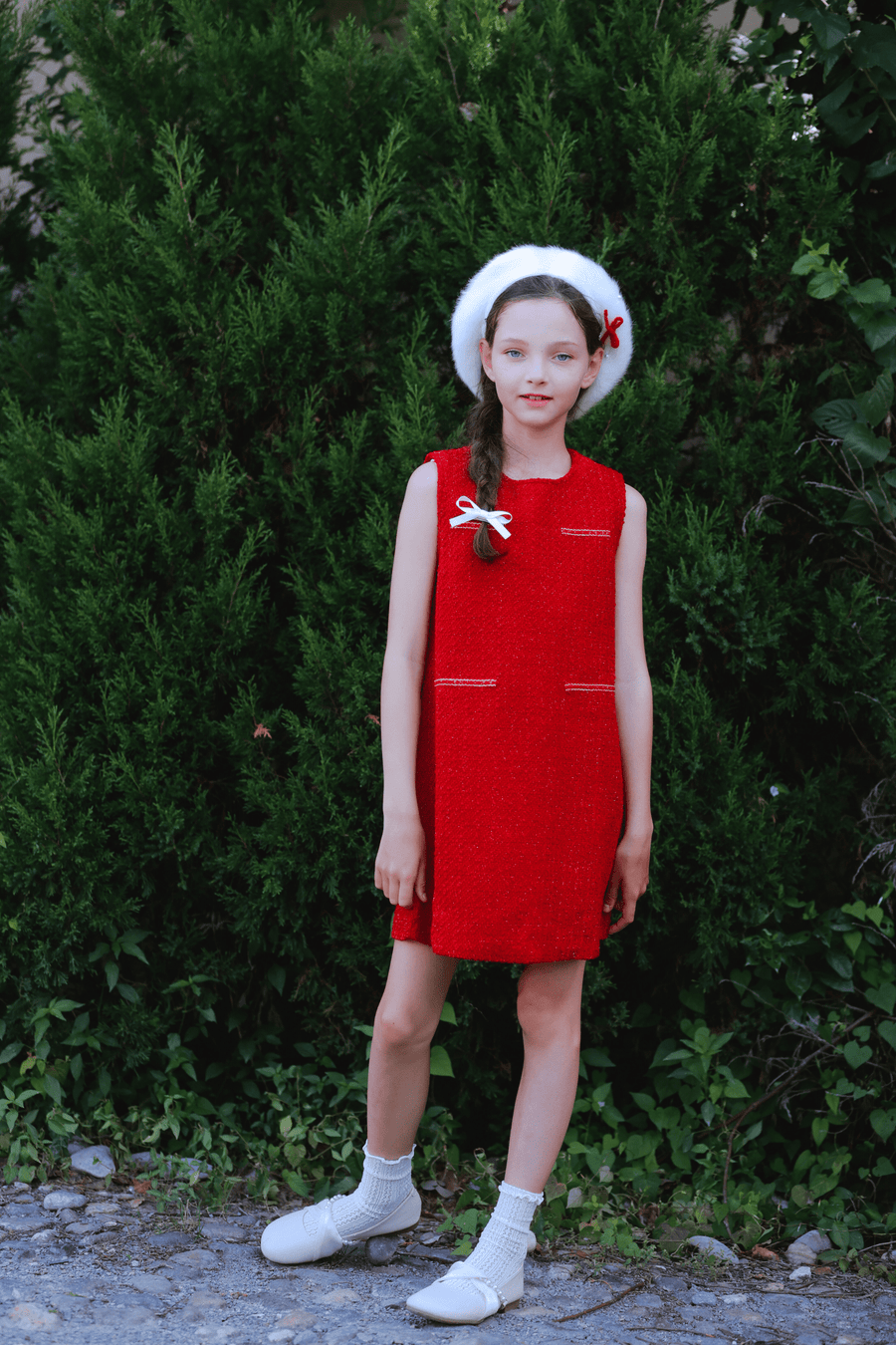 A young girl stands outdoors in front of green bushes, wearing the KIKI and JOJO Girls Tweed Dress in Cranberry Red, paired with white shoes, socks, and a beret with a red bow—ideal for festive occasions.