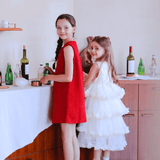 Two young girls stand by a wooden sideboard with drinks and glassware; one wears the KIKI and JOJO Girls Tweed Dress in cranberry red, the other a white ruffled dress—perfect festive attire—as they both look toward the camera.