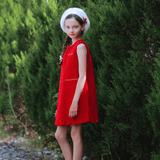 A young girl wearing the KIKI and JOJO Girls Tweed Dress in Cranberry Red with a white beret and shoes stands on a stone path beside tall green bushes.