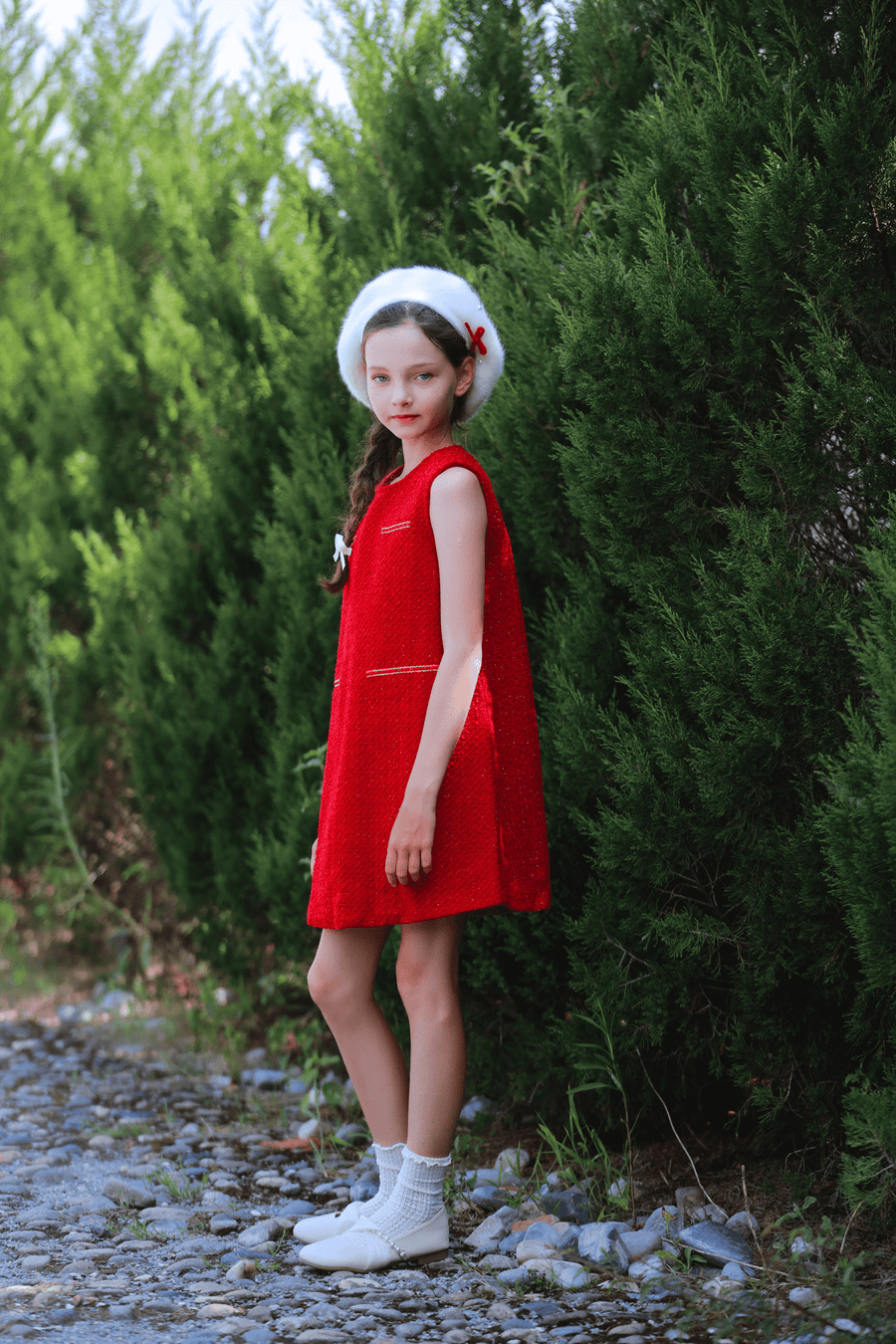 A young girl wearing the KIKI and JOJO Girls Tweed Dress in Cranberry Red with a white beret and shoes stands on a stone path beside tall green bushes.