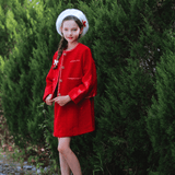 A young girl wears the KIKI and JOJO Girls Tweed Dress in cranberry red with a sleeveless A-line silhouette, white socks, shoes, and hat, standing on a gravel path before tall green bushes—perfect for festive occasions.
