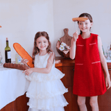 Indoors, two young girls celebrate: one in a white dress holds baguettes, while the other smiles in the KIKI and JOJO Girls Tweed Dress - Cranberry Red, holding bread on a stick; bottles and bread sit on the table behind them.