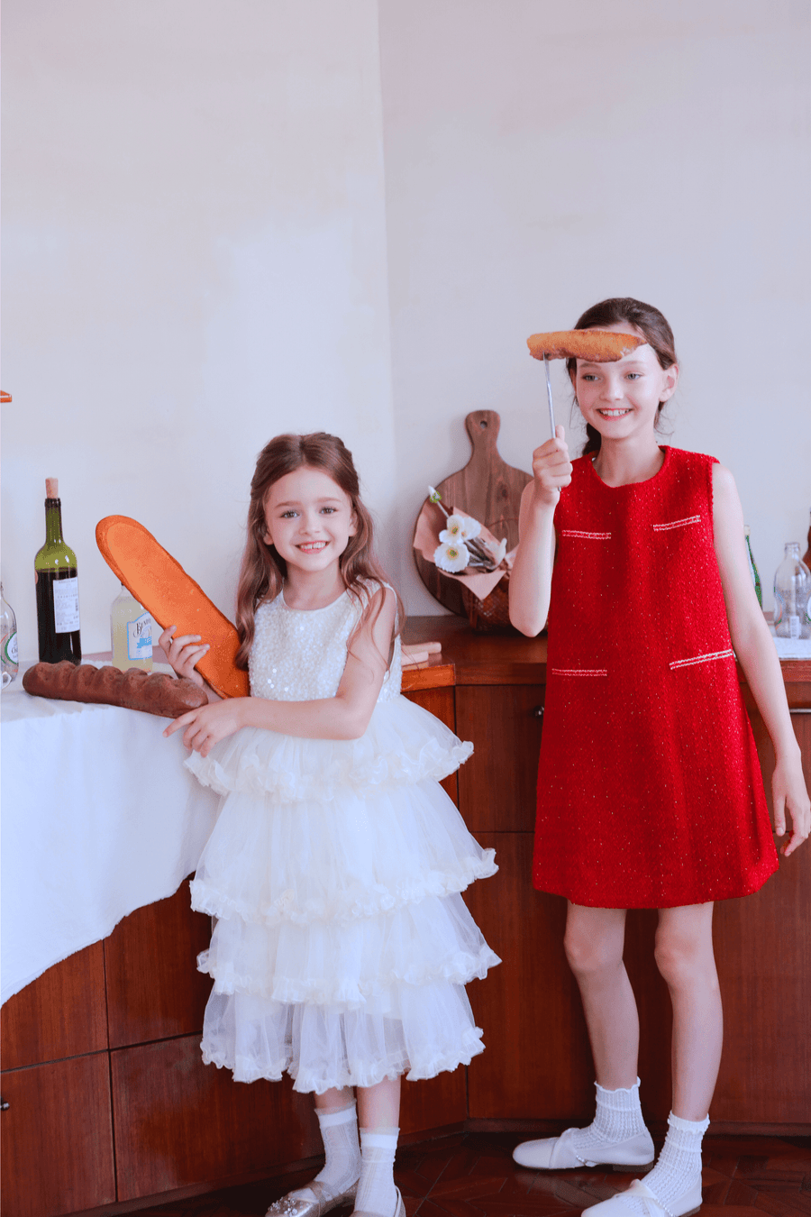 Indoors, two young girls celebrate: one in a white dress holds baguettes, while the other smiles in the KIKI and JOJO Girls Tweed Dress - Cranberry Red, holding bread on a stick; bottles and bread sit on the table behind them.