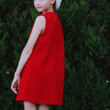 A young girl in the KIKI and JOJO Girls Tweed Dress - Cranberry Red, a sleeveless A-line style, pairs it with a white fur hat and poses outdoors by green bushes—an ideal look for festive occasions.