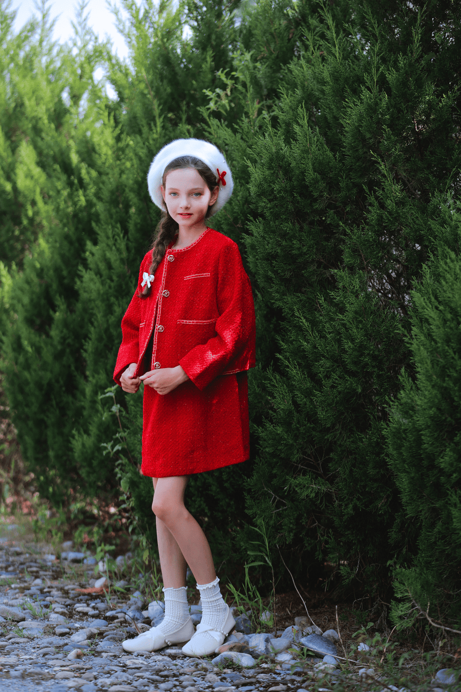 A girl in the KIKI and JOJO Girls Tweed Jacket - Cranberry Red, with champagne gold buttons, white socks and shoes, and a white beret stands on a stone path before tall green bushes.