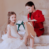 Two young girls sit on stairs; one smiles in a white dress while the other opens a brown gift bag, wearing the KIKI and JOJO Girls Tweed Jacket in Cranberry Red. Wrapped ribboned presents are visible in the background.