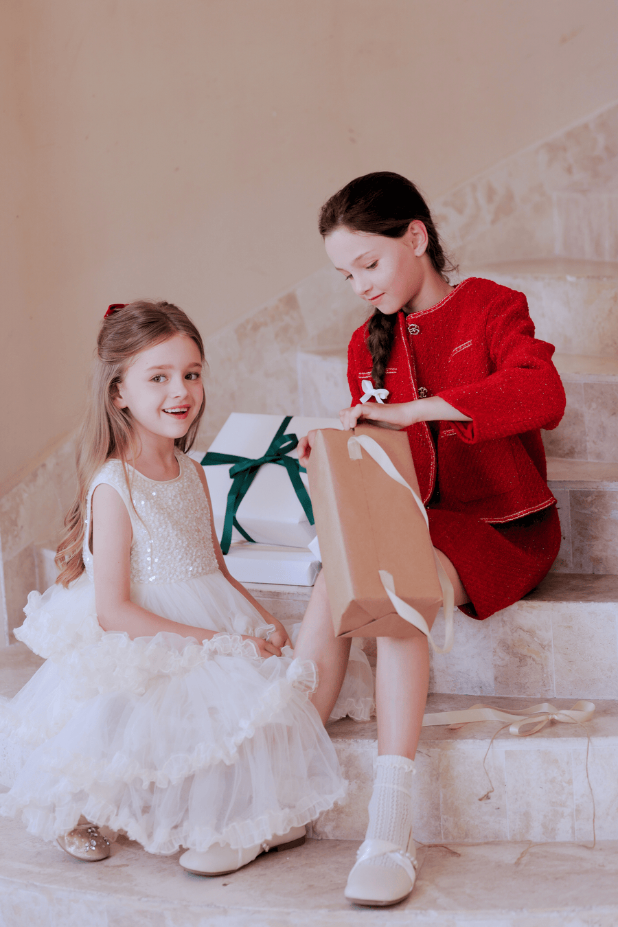 Two young girls sit on stairs; one smiles in a white dress while the other opens a brown gift bag, wearing the KIKI and JOJO Girls Tweed Jacket in Cranberry Red. Wrapped ribboned presents are visible in the background.
