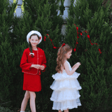 Two young girls stand outside by green bushes; one wears the KIKI and JOJO Girls Tweed Jacket in Cranberry Red and a white hat, while the other has a layered white dress with her brown hair in a ponytail.