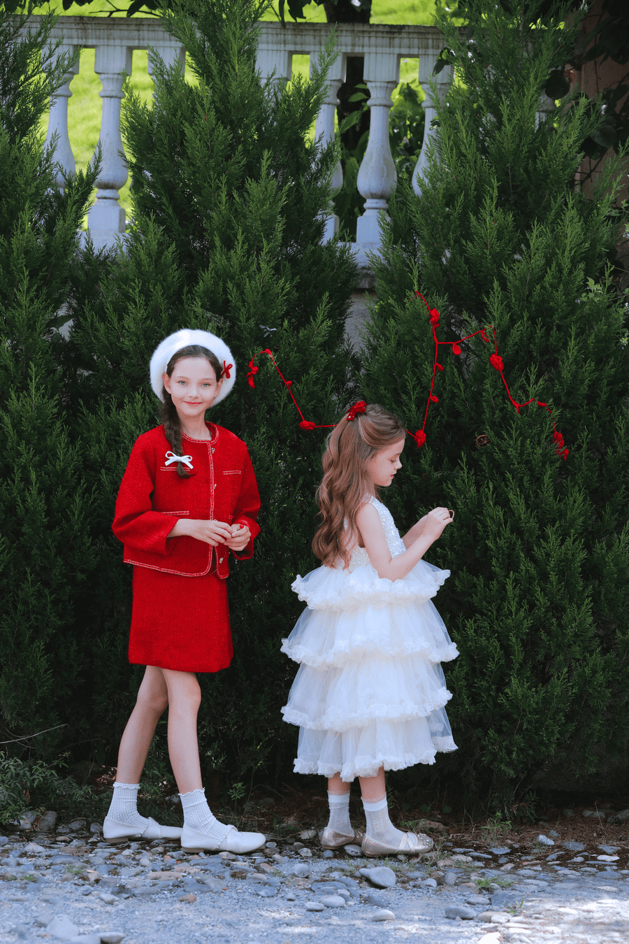 Two young girls stand outside by green bushes; one wears the KIKI and JOJO Girls Tweed Jacket in Cranberry Red and a white hat, while the other has a layered white dress with her brown hair in a ponytail.