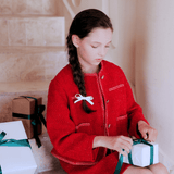 A girl with a braid, wearing the KIKI and JOJO Girls Tweed Jacket in Cranberry Red with champagne gold buttons, sits on stairs untying the ribbon on a gift box, with other wrapped presents next to her.