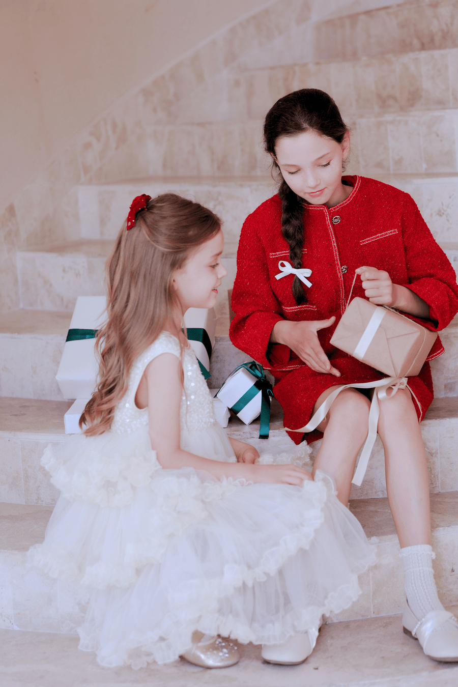 Two young girls sit on stairs with wrapped gifts; one wears the KIKI and JOJO Girls Tweed Jacket in Cranberry Red, holding a present, while the other in white with champagne gold buttons looks on.